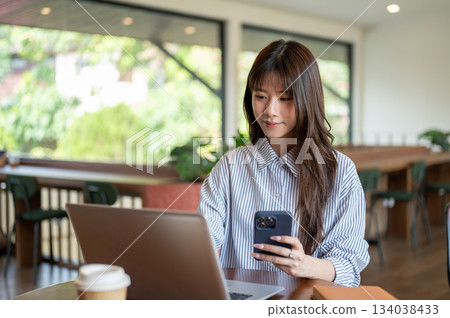 Asian woman in blue shirt holding phone and looking at laptop while sitting at wooden table in cafe. Asian woman in blue shirt holding phone and looking at laptop while sitting at wooden table in cafe. 134038433