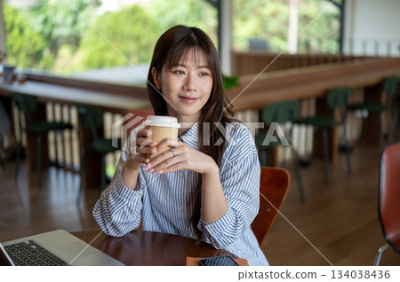 Smiling asian woman holding coffee cup with both hand while sitting with laptop at cafe wooden table Smiling asian woman holding coffee cup with both hand while sitting with laptop at cafe wooden table 134038436