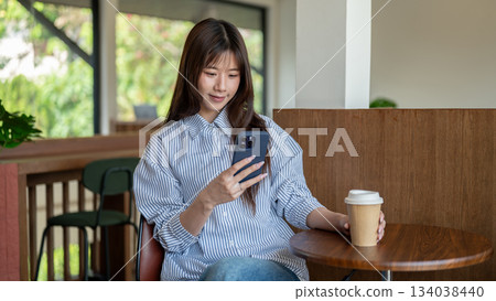 Smiling asian woman looking at phone and holding coffee while sitting at wooden stool table in cafe. Smiling asian woman looking at phone and holding coffee while sitting at wooden stool table in cafe. 134038440