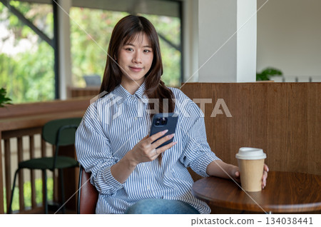 Smiling asian woman in blue shirt holding phone and coffee cup while sitting at wooden table in cafe Smiling asian woman in blue shirt holding phone and coffee cup while sitting at wooden table in cafe 134038441