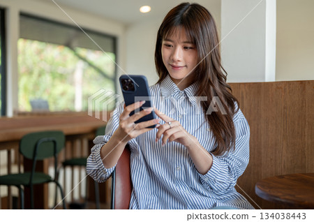 Smiling asian woman in blue shirt looking and typing on her phone while sitting at cafe wooden table 134038443