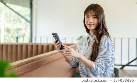 Smiling asian woman holding phone and lookin away while sitting at wooden counter in cafe or library 134038450