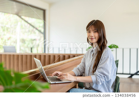 Smiling asian woman is typing on laptop with both hand sitting at wooden counter in cafe or library. 134038456