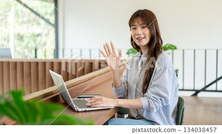 Blue shirt asian woman smiling and waving a hand while sitting at wooden counter in cafe or library Blue shirt asian woman smiling and waving a hand while sitting at wooden counter in cafe or library 134038457