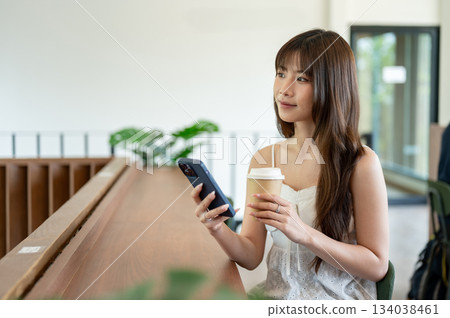 Asian woman in camisole holding coffee and looking away from phone while sitting at wooden counter. Asian woman in camisole holding coffee and looking away from phone while sitting at wooden counter. 134038461