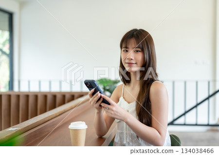 Asian woman in camisole smiling with smartphone in her hands while sitting at cafe's wooden counter. Asian woman in camisole smiling with smartphone in her hands while sitting at cafe's wooden counter. 134038466