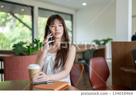 Asian woman in camisole talking on the phone and grabbing coffee while sitting at cafe wooden table. 134038469