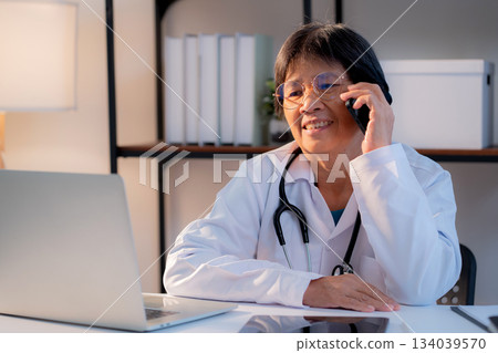 Senior asian doctor speak on smartphone while working on laptop in medical office. 134039570