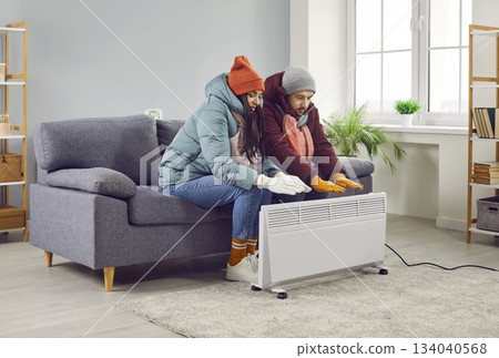 Young frozen couple sitting at home and trying to warm their hands on electric heater. Young frozen couple sitting at home and trying to warm their hands on electric heater. 134040568