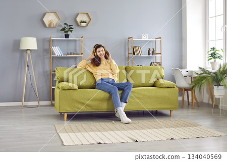 Young smiling woman sitting on the sofa at home and listening to music with headphones. Young smiling woman sitting on the sofa at home and listening to music with headphones. 134040569