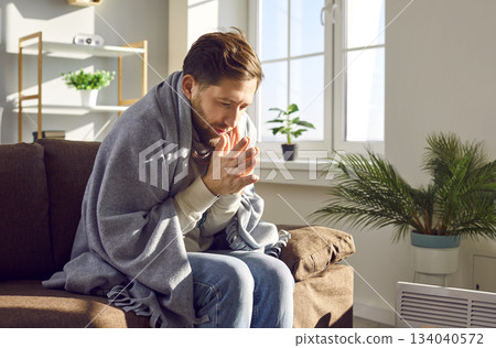 Young frozen man sitting wrapped in blanket at home near the electric heater. 134040572