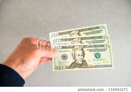 左手拿著三張20美元鈔票，從左側展示的斜角照片。 134041071