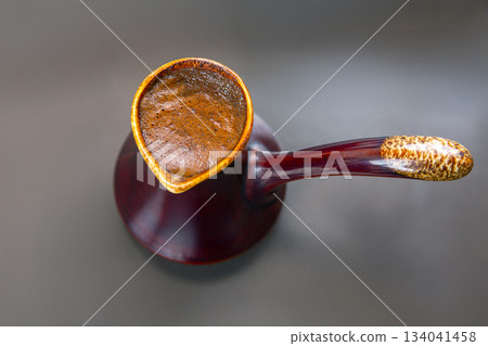 Close-up of a shiny cezve filled with frothy Turkish coffee on a sleek gray surface. 134041458