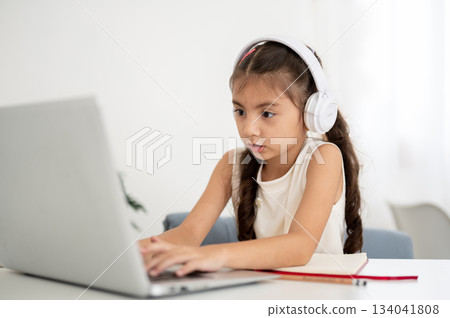 Asian girl kid wearing headphones looking and typing on laptop while sitting at white table in house Asian girl kid wearing headphones looking and typing on laptop while sitting at white table in house 134041808