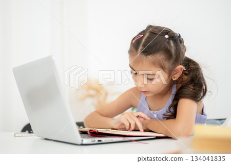 Asian kid girl reading or looking at an open notebook over a laptop while sitting at white table in house. Asian kid girl reading or looking at an open notebook over a laptop while sitting at white table in house. 134041835