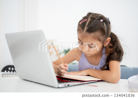 Asian kid girl looking down at an open notebook over a laptop while sitting at white table in house. Asian kid girl looking down at an open notebook over a laptop while sitting at white table in house. 134041836