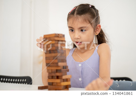 Asian kid or little girl in purple shirt playing wooden tower building game on table in white room Asian kid or little girl in purple shirt playing wooden tower building game on table in white room 134041861