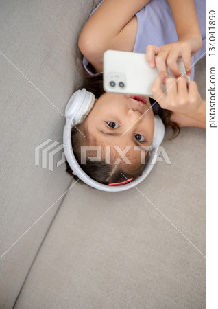 Asian kid or a little girl with headphones holding phone with both hands while lying on fabric sofa. 134041890