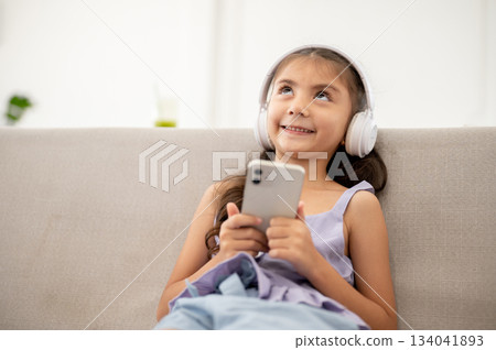 Asian kid or girl with headphone holding smartphone and smiling funnily while sitting on fabric sofa Asian kid or girl with headphone holding smartphone and smiling funnily while sitting on fabric sofa 134041893
