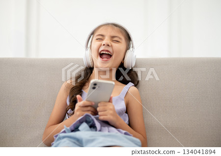 Asian kid or a girl with white headphones is holding phone and laughing while sitting on fabric sofa 134041894