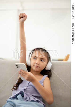An asian kid or girl with white headphones holding phone and raising one arm while sitting on sofa. 134041896