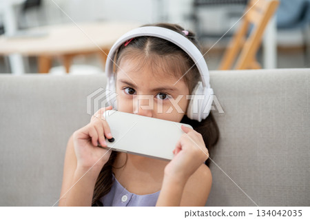Asian kid or girl with white headphones holding smartphone covered her mouth while sitting on sofa. 134042035