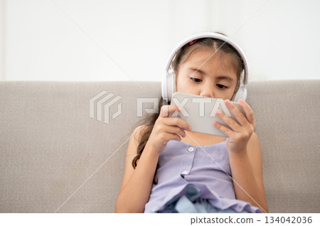Asian kid or girl with white headphones holding smartphone and looking closely while sitting on sofa 134042036
