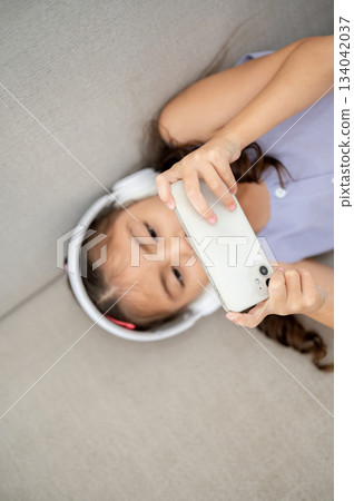 Asian kid or a girl wearing white headphones holding smartphone with both hands while lying on sofa 134042037