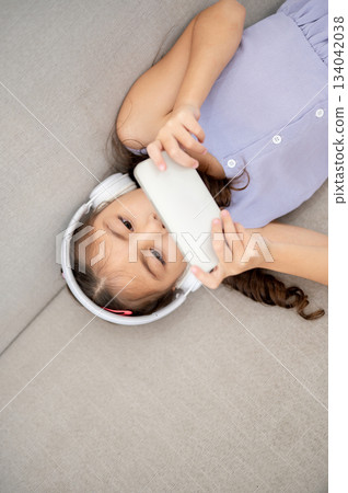 Asian kid or a girl wearing white headphones holding smartphone in both hands while lying on sofa 134042038