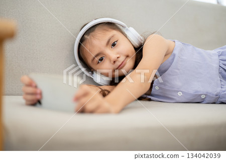 Asian kid or a girl wearing white headphones holding phone while lying facing camera on fabric sofa. Asian kid or a girl wearing white headphones holding phone while lying facing camera on fabric sofa. 134042039