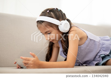 An asian kid or a girl wearing white headphones looking at her smartphone while lying on fabric sofa An asian kid or a girl wearing white headphones looking at her smartphone while lying on fabric sofa 134042040
