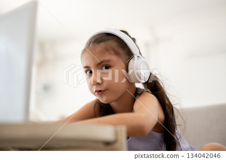 Asian kid or a girl wearing headphones giving a funny look while sitting with laptop on white table. 134042046