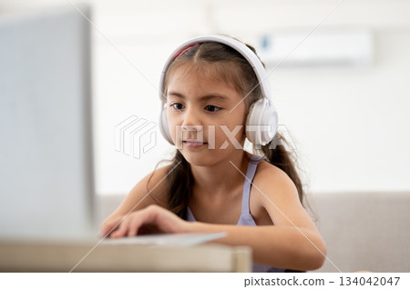 Asian kid or a girl wearing headphones is staring at her laptop on white table while sitting on sofa 134042047