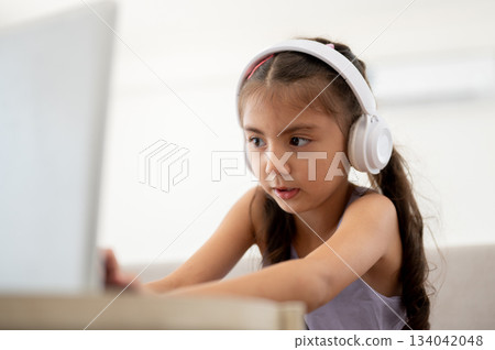 Asian kid or a girl with headphones looking curiously at laptop on white table while sitting on sofa 134042048