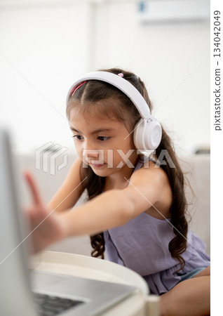 Asian kid or girl wearing headphones touching on laptop screen at white table while sitting on sofa. 134042049