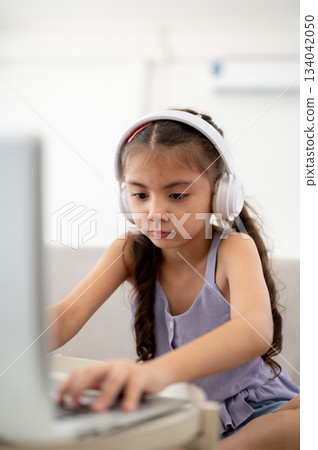 An asian kid or girl wearing headphones typing on her laptop at white table while sitting on sofa. 134042050