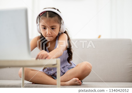 Asian kid or girl wearing headphone typing on laptop with clenched lip while sitting on frabic sofa. Asian kid or girl wearing headphone typing on laptop with clenched lip while sitting on frabic sofa. 134042052