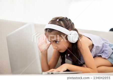 Asian kid or girl with headphone looking at laptop and raising a hand while crouching on fabric sofa 134042055