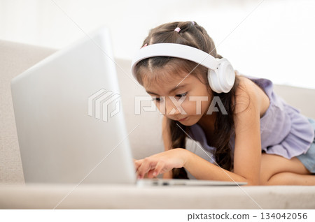 Asian kid or a girl wearing white headphones is looking at her laptop while crouching on fabric sofa 134042056
