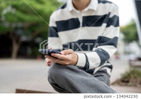 Close up of a man holding smartphone while sitting cross legged on a wooden bench in the parking lot 134042230