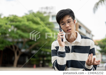 Asian man wearing glasses is talking on smartphone with explaining hand gesture in parking lot. 134042232
