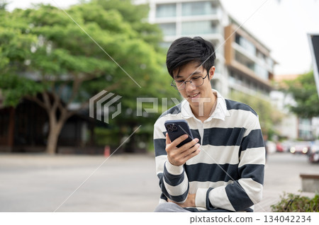 Asian man wearing glasses looking at smartphone while sitting on a wooden bench in the parking lot. Asian man wearing glasses looking at smartphone while sitting on a wooden bench in the parking lot. 134042234