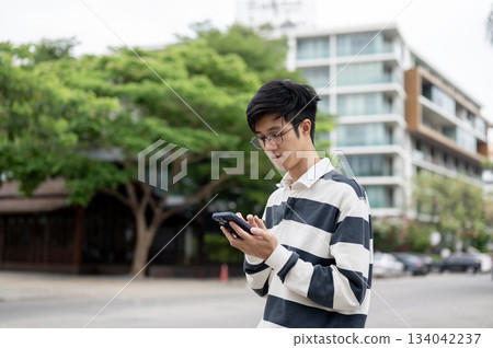 Asian man wearing glasses is looking and typing at his smartphone while standing in the parking lot. 134042237