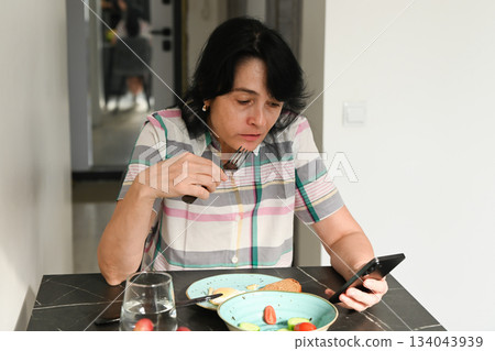 A 50 year old Russian woman eats while checking her phone, showing distracted eating and a growing dependence on constant phone use during meals. 134043939