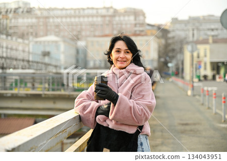 A Russian woman 50 years old smiles on a city bridge holding a warm coffee cup in soft spring light feeling calm awareness gentle energy and a sense of morning freedom 134043951