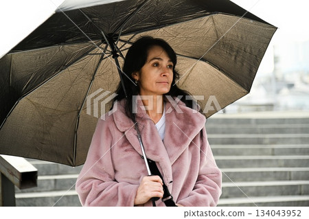 Russian woman about 50 years old stands outdoors under an umbrella on city steps looking thoughtful and calm in cool weather Russian woman about 50 years old stands outdoors under an umbrella on city steps looking thoughtful and calm in cool weather 134043952