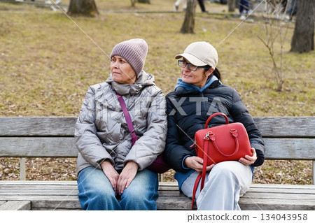 Two Russian friends 50 and 60 years old sit in a spring park looking left enjoying a quiet moment together with calm presence freedom and relaxed connection. Two Russian friends 50 and 60 years old sit in a spring park looking left enjoying a quiet moment together with calm presence freedom and relaxed connection. 134043958