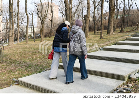 Two Russian friends 50 and 60 years old walk arm in arm up the park stairs in spring enjoying calm movement shared presence and a quiet mindful moment together. Two Russian friends 50 and 60 years old walk arm in arm up the park stairs in spring enjoying calm movement shared presence and a quiet mindful moment together. 134043959