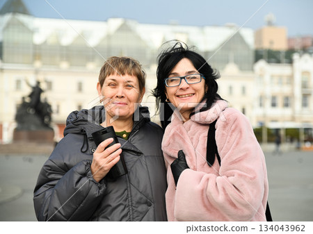 Two Russian friends, 50 and 60 years old, smiling warmly at the camera in the city, sharing joy, confidence, lightness, and the ease of mature female friendship. Two Russian friends, 50 and 60 years old, smiling warmly at the camera in the city, sharing joy, confidence, lightness, and the ease of mature female friendship. 134043962