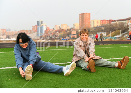 Two Russian women aged 50 and 60 stretch forward while sitting on grass at a stadium in Vladivostok, staying active together and supporting healthy life 134043964